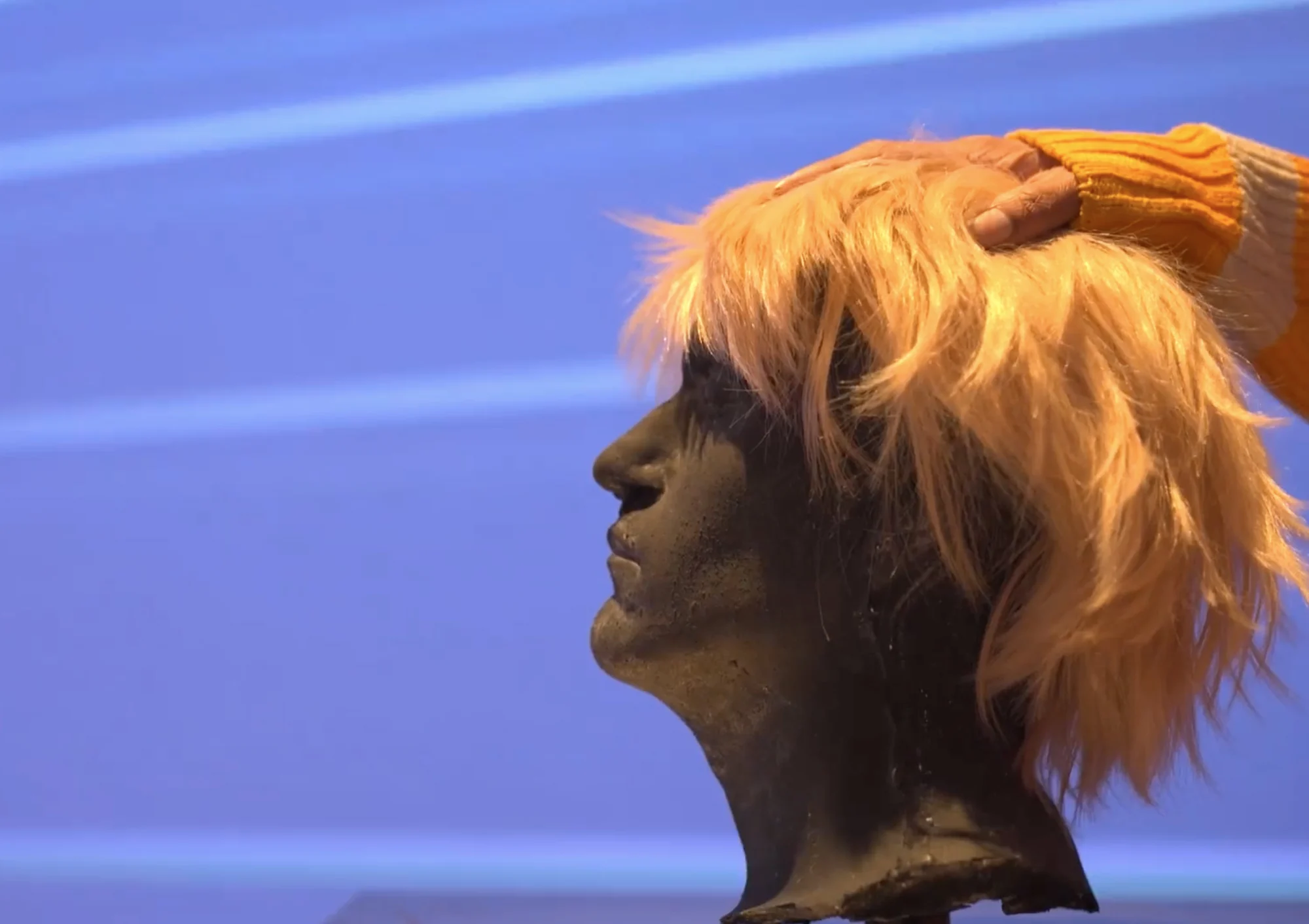 A sculptural head with orange hair against the blue projection of It’s Just a Dream
